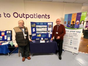 Two smiling gentleman stand in YDH outpatients corridor in front of a banner and a table introducing the trust governors. 
