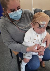 A child sitting still on their mother's lap with a device to measure the response to sound is attached to the child's head