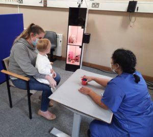 A child completing a hearing test to measure their minimum audible level. The child is sat in front of a box with their head turned towards the object in the box. 