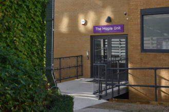 An image of the Maple Unit in Yeovil Hospital. In the background is the entrance of the ward which has wide glass doors and a purple building sign above the doors. In the foreground is some foliage on the left side of the image.
