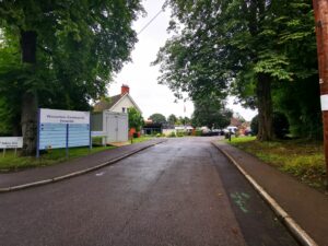 An image of the entrance to the site at Wincanton. It is a large road with big very old trees either side. To the left is a sign with instructions on where visitors need to go with a faint sight of a building in the back gorund.