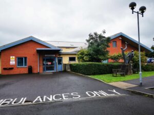 West Mendip Hospital is a building that has two pitched roofs on either side with a small flat roofed building in the middle. These are all attached. The front of the pitched roof have blue edging and the rest is red brick. The middle of the building is off white.