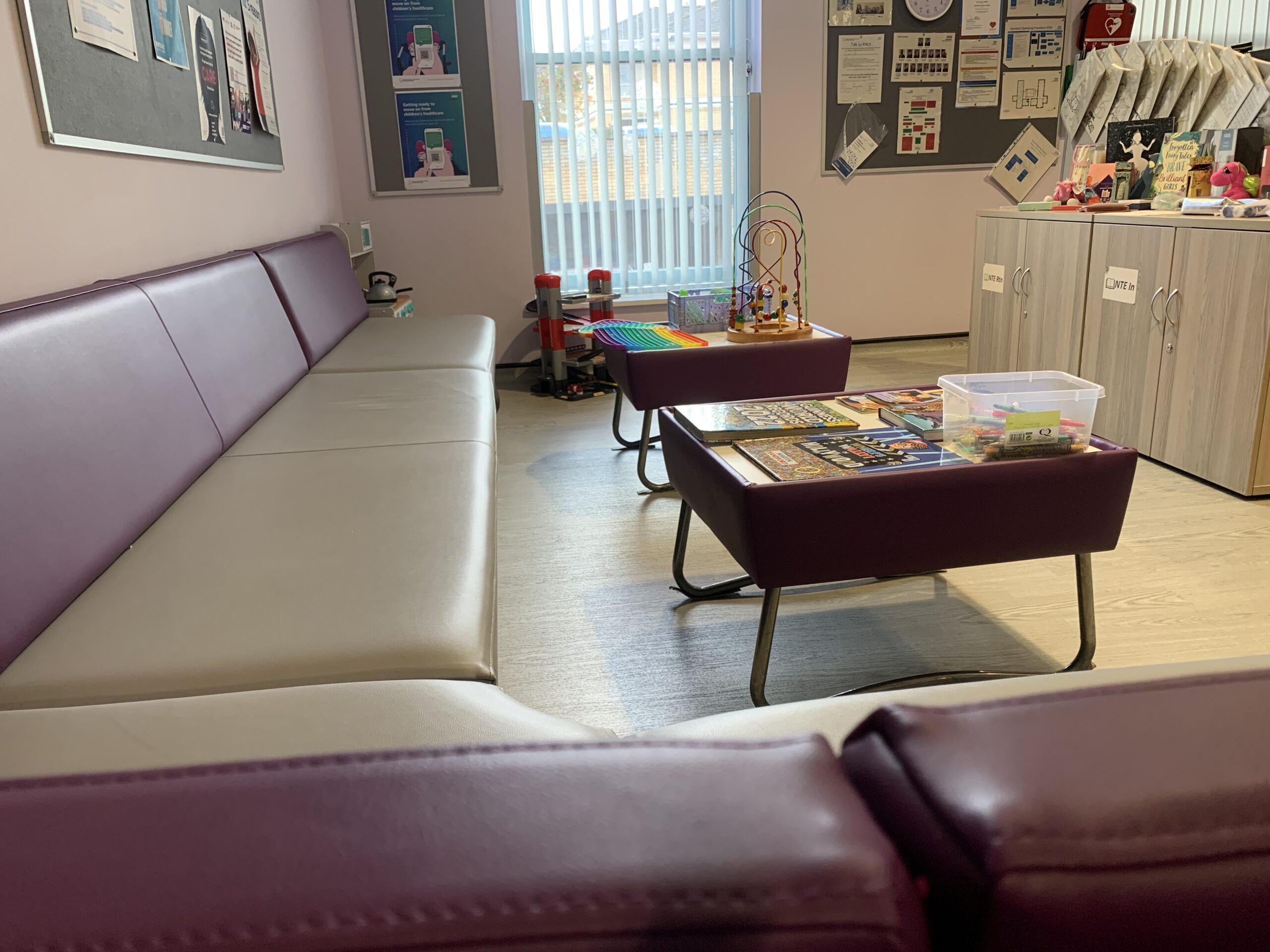 An image of the inside of the waiting room of the Community families hub. the waiting room has a purple and grey corner sofa, nearby are two small low coffee tables with various toys, children books and a clear box filled with various colouring pencils. In the background are various posters and information pinned to the wall.