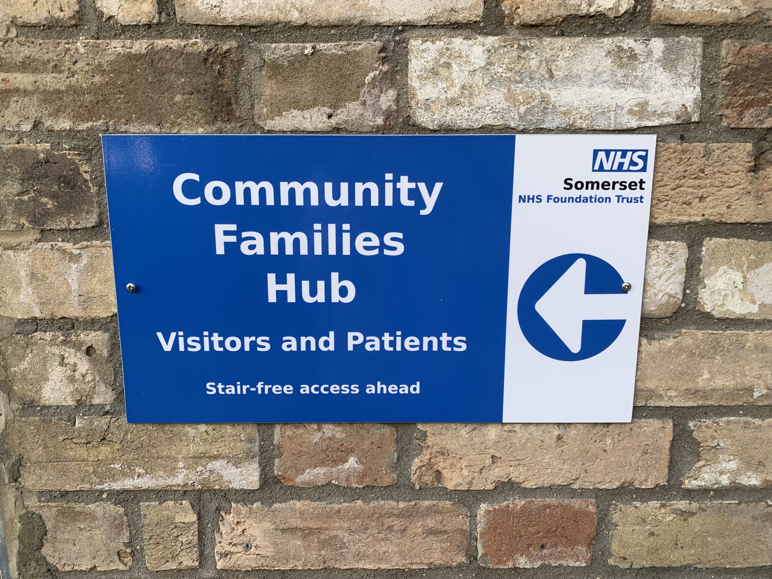 A blue and white building sign with a arrow sign that reads: NHS Somerset NHS Foundation Trust, Community Families Hub Visitors and Patients Stair-free access ahead.