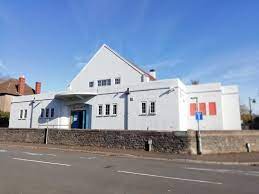 An image of a modern white building with a pitched roof, blue door and multiple small windows. A stone walls runs along the front of the building.