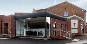 A modern church building with glass extension at the front and a cross symbol on the right side of the building.