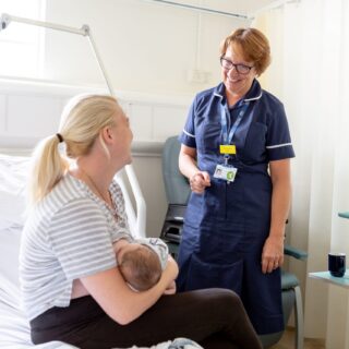 A parent holding a baby in their arms with a NHS colleague standing next to them having a happy conversation.