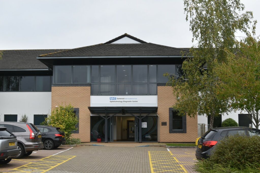 An image of Harrison House. In the background is an image of a building entrance with windows on the first floor reaching all around the building. The entrance has a white NHS building sign with the name of the building and NHS logo. In the foreground is the parking area of the building which has several cars parked near the building and yellow parking lines on the floor.