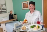 A lady in chef whites and a navy chef's hat stands smiling at the front of the image holding a tray with red fruit juice in a glass, a plate with white meat and green beans and brocolli with a side of new potatoes and a fruit salad dessert. Behind the chef's shoulder to the right is a wooden door and white frame and to the left is a neatly made hospital bed with table over the centre and a patient in navy blue pyjamas sits perched on the side of the bed at the pillow end with a light wood effect side table behind to the side. The walls are white and one teal which also features framed artwork that is unclear in the background.