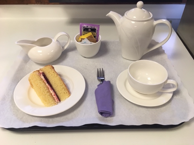 A tray with a white teapot, cup and saucer, small milk jug, a cup with a sugar packets inside, a plate with a slice of Victorian sponge cake and a fork with a purple napkin wrapped around it. The tray is lined with a paper doily.