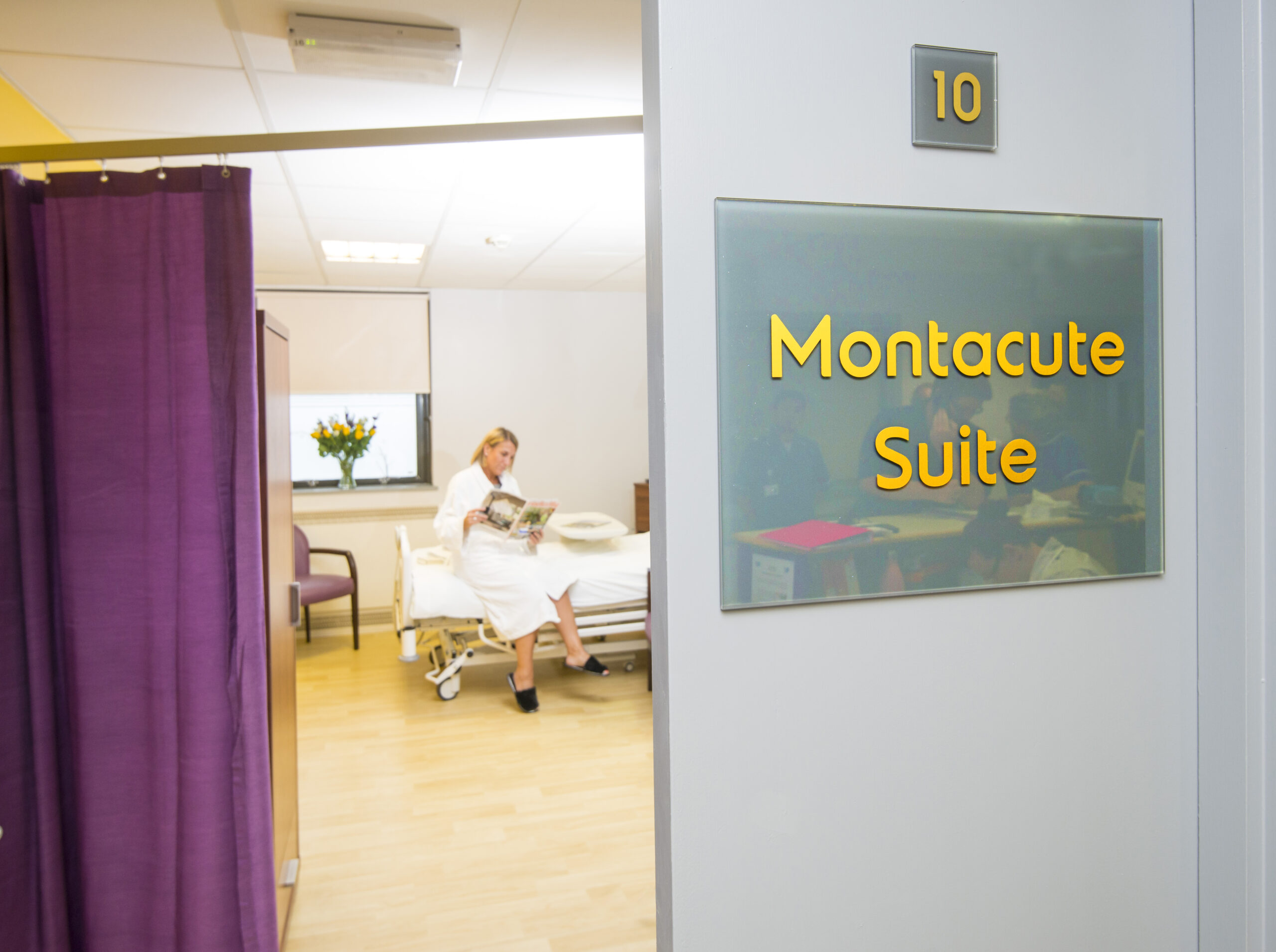 Entrance to a hospital room labelled “Montacute Suite,” room 10. A person is sitting on a hospital bed inside, reading. A purple curtain partially covers the doorway, and flowers are visible on a table near the window.