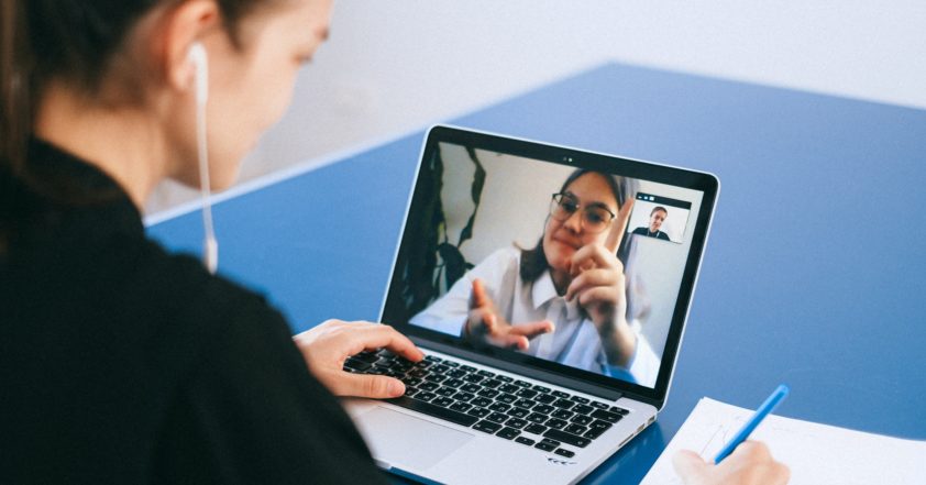 A person looking at a laptop which has an image of another person. They look like they are in conversation. The person looking at the laptop has a headphone in her right ear, left hand on the keyboard and right hand has a pen writing on the paper. the Table used is blue with white edging.