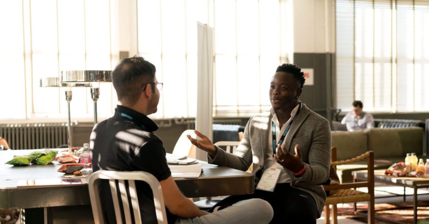 Image of two people facing each other in conversation. Both dressed really smart. The setting is almost a break room with lots of tables and chairs.