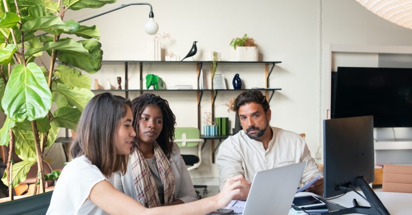 Three people all looking at a laptop screen and another monitor. They are sat at a white round table pointing at the screen. In the background is some bookshelf's with supports from trees sticks. There is banana plant on the left hand side of the image.