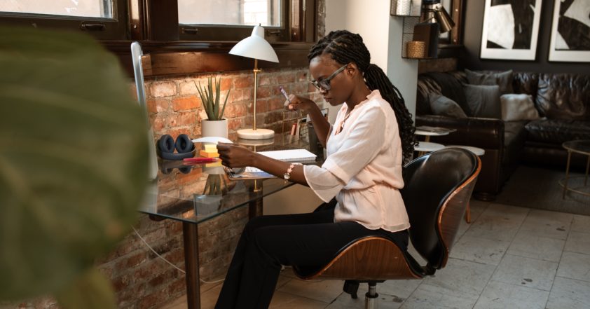 An image of a person working at a busy but clean desk. Th person looks relaxed in her work. The room setting is almost a factory/ industrial setting with brick walls. There is black and white abstract paintings in the very distance of the image.