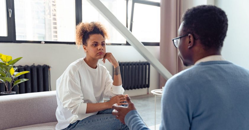 Image is of two people. one is talking to the other that is listening. The person listening is sat on a grey comfortable sofa which is in front of some old Victorian radiators which are below some very big windows which makes the room very light. There is a supporting roof pole that goes across the window.