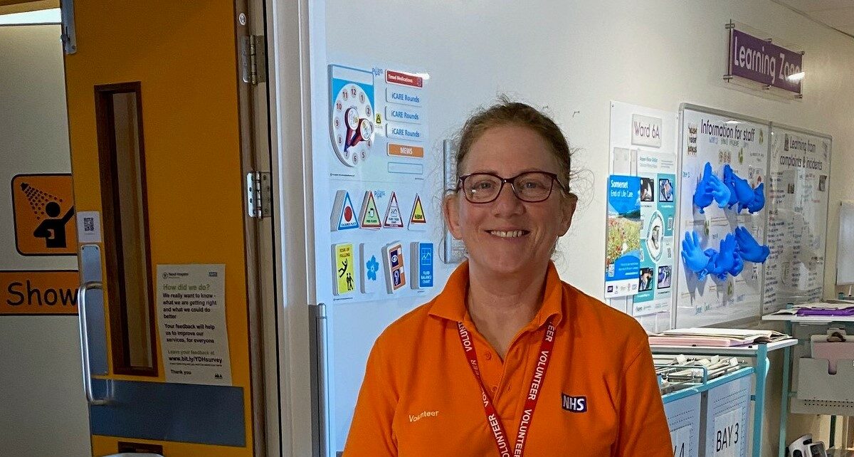 Jane is stood behind a cleaning trolley on a ward. She is stood infront of some double doors which is next to a wall of lots of baords and posters.