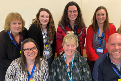 maternal mental health service team members are photographed with the heads, shoulders and tops of torsos of three colleagues in the front row and five further colleagues gathered behind with the tallest in the centre. Each of the team is wearing daytime clothing and blue lanyards around their necks, smiling directly towards the camera.