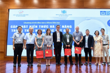 Pictured with the Vietnam team are: (3rd from left) Dr Joanna Brown - consultant radiologist, and (5th from left) Dr Andy Lowe. They are all stood infront of a large projected presentation, which is written in Vietnamese.