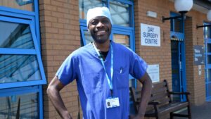 Paul Ennis, a healthcare assistant in our Day Surgery Centre at Musgrove Park Hospital who is stood in front of the Day Surgery Centre dressed in blue scrubs.