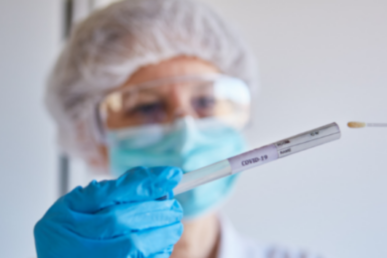 A photo of a woman wearing a clinical hat, glasses and gloves, holding a COVID-19 test.