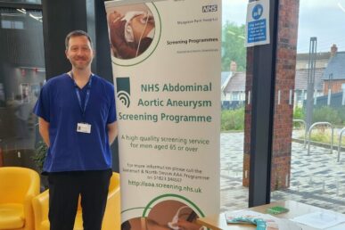 A photo of an abdominal aortic aneurysm screening practitioner stood next to a banner advertising screening sessions.
