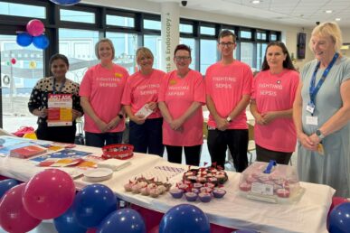Seven NHS colleagues from the sepsis team stood next to a table with Sepsis-related items on. Five of the people are wearing pink t-shirts.