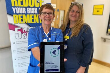 Two colleagues from Somerset NHS Foundation Trust's research team standing indoors. The person on the left is wearing a blue uniform with an NHS logo, and the person on the right is wearing a dark outfit with a yellow badge. Both individuals are holding a tablet device, displaying the Five Lives MED website.