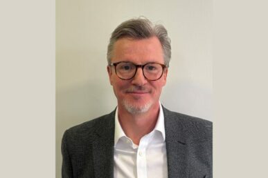 Professional headshot of a person wearing round glasses, a grey blazer and white shirt, looking at the camera against a plain light-coloured background.