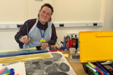 A photo of art psychotherapist Sevadeva Hamlin holding a paintbrush with lots of arts and crafts materials on the table.
