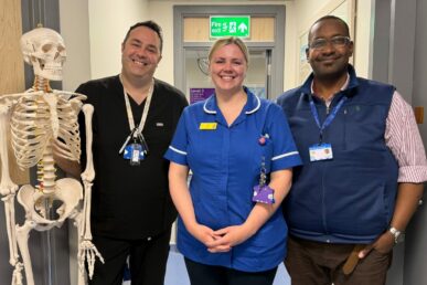 A photo of three clinicians involved in the spinal service stood in a corridor next to a model skeleton.